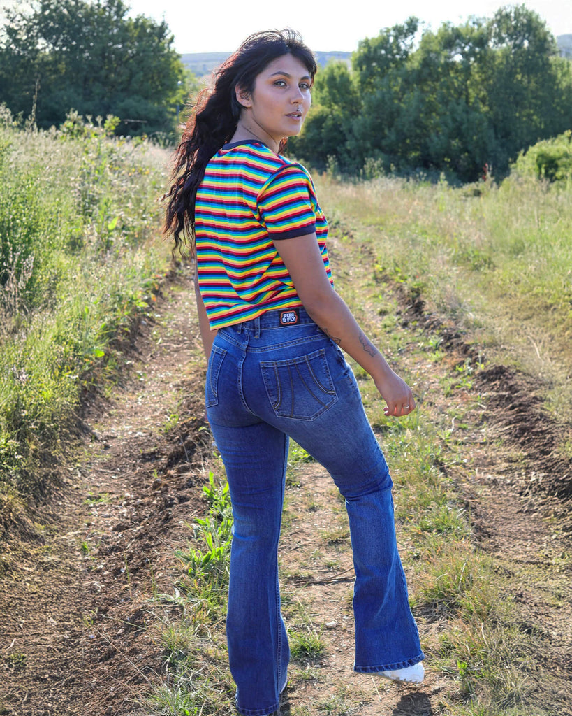 Meera is stood in a grassy field wearing the stone wash blue distress stretch denim rock n roll bell bottom flares with a retro rainbow short sleeve tshirt and white trainers. She is facing away with one leg bent forward to highlight the jean shape and looking over her shoulder to camera.