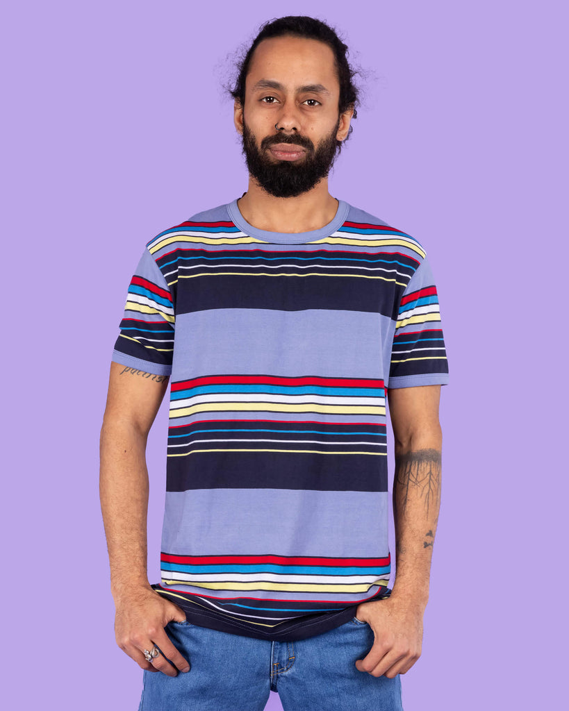 Richard, a hispanic male model with dark hair in a bun and a beard, is stood in a photography studio in Hove in front of a lilac backdrop wearing Vintage Blue Stripe Tee with blue jeans. The blue t shirt has horizontal lines in red, blue, white, yellow and navy. Richard is posing to the camera with his thumbs resting in the front pockets of his jeans and the photo is cropped at the thighs.