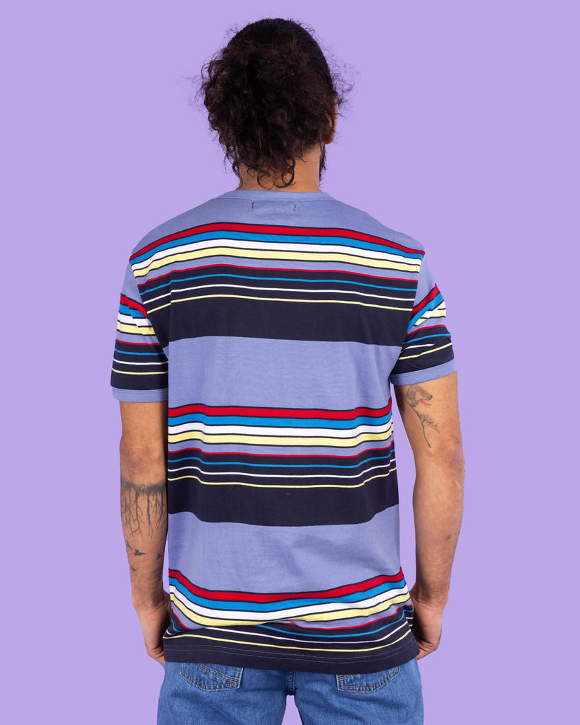 Richard, a hispanic male model with dark hair in a bun and a beard, is stood in a photography studio in Hove in front of a lilac backdrop wearing Vintage Blue Stripe Tee with blue jeans. The blue t shirt has horizontal lines in red, blue, white, yellow and navy. Richard is stood with his back to the camera and arms down by his sides.