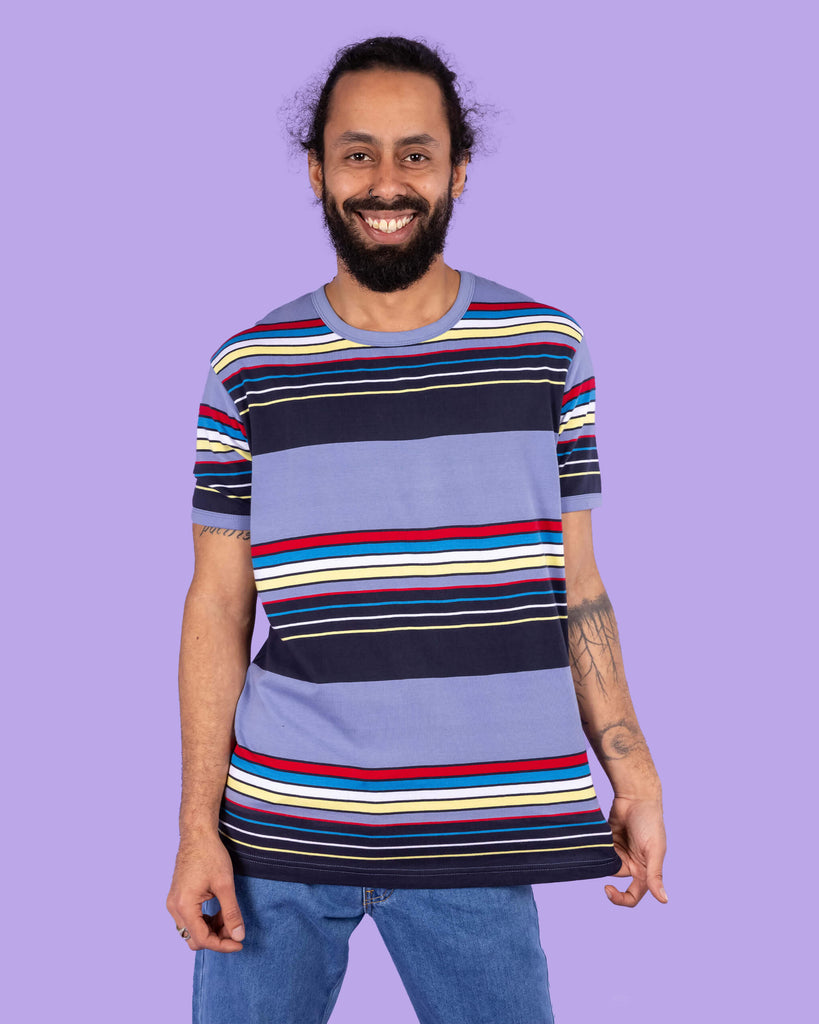 Richard, a hispanic male model with dark hair in a bun and a beard, is stood in a photography studio in Hove in front of a lilac backdrop wearing Vintage Blue Stripe Tee with blue jeans. The blue t shirt has horizontal lines in red, blue, white, yellow and navy. Richard is facing the camera smiling and is holding the bottom of the t shirt with one hand and other resting down by his side. The photo is cropped at the thighs.