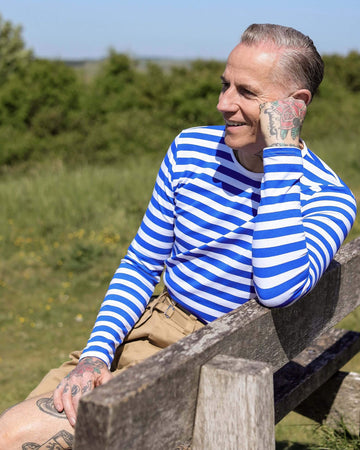 Jim is sitting in a park on a bench, his face resting on his hand whilst wearing tan shorts and the Royal & White Stripe Long Sleeve T Shirt