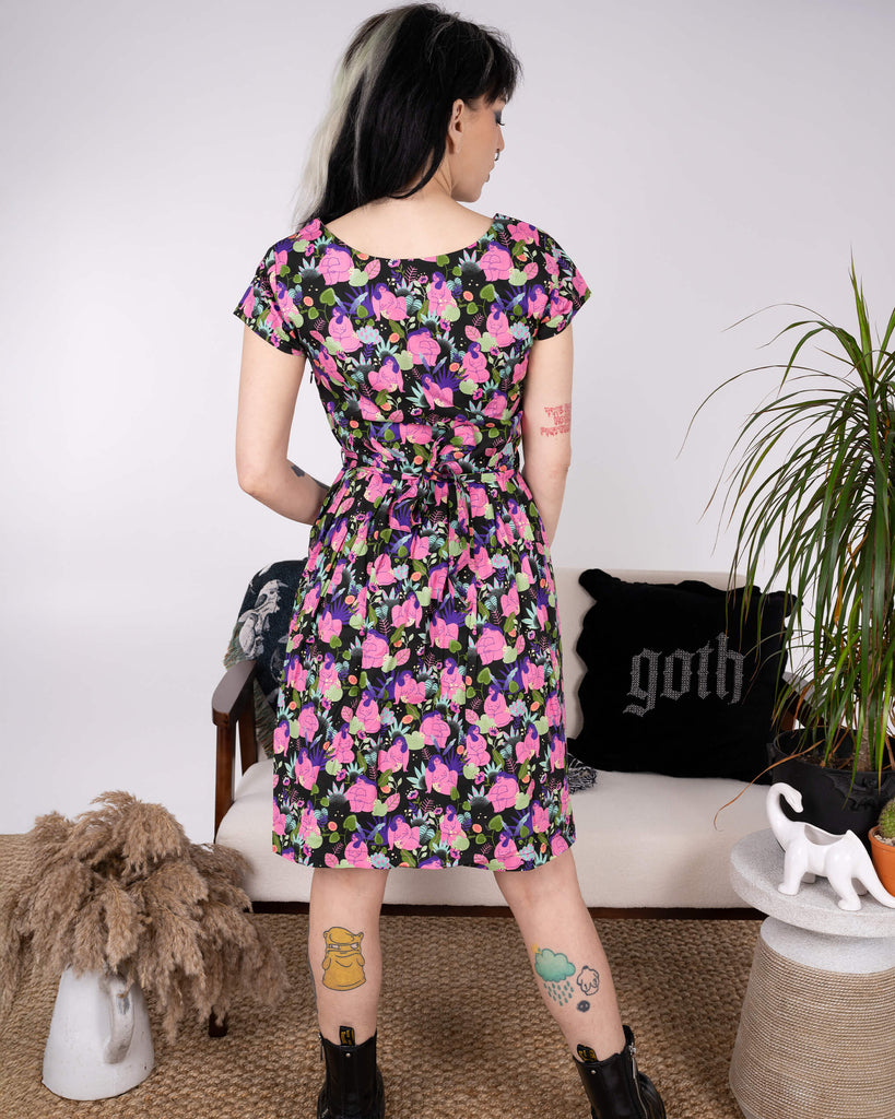 Back view of the Run & Fly x Marie Boiseau Bodies Stretch Belted Tea Dress with Pockets. The model highlights the dress's fit, with a tied belt accentuating the waist. The black background contrasts the bright, artistic print, crafted by a small ethical slow fashion brand.