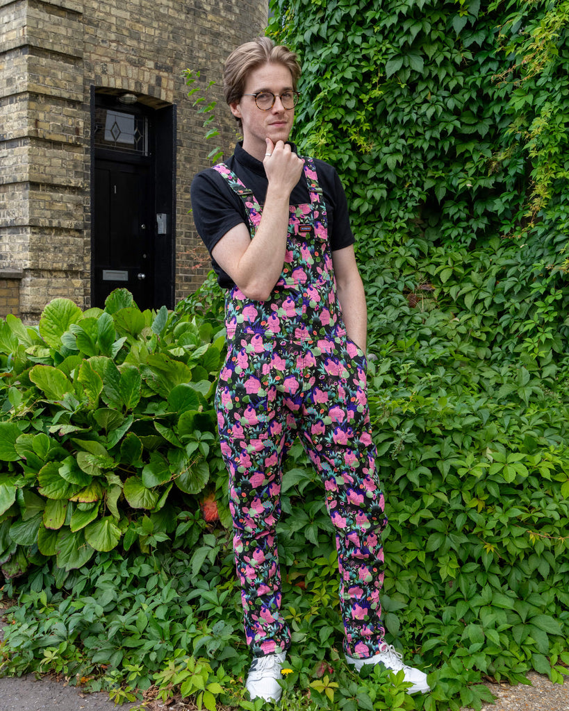 Model Jack Roper wears vibrant Run & Fly stretch twill dungarees featuring a fun 'Bodies' print by marie boiseau. The colourful pattern showcases pink bodies with green florals and fun details, paired with a black polo shirt. Set against lush greenery, this ethical slow fashion piece celebrates unique, sustainable style. Perfect for lovers of playful, bold fashion. Run & Fly is a small UK-based brand known for ethical, eco-conscious clothing.