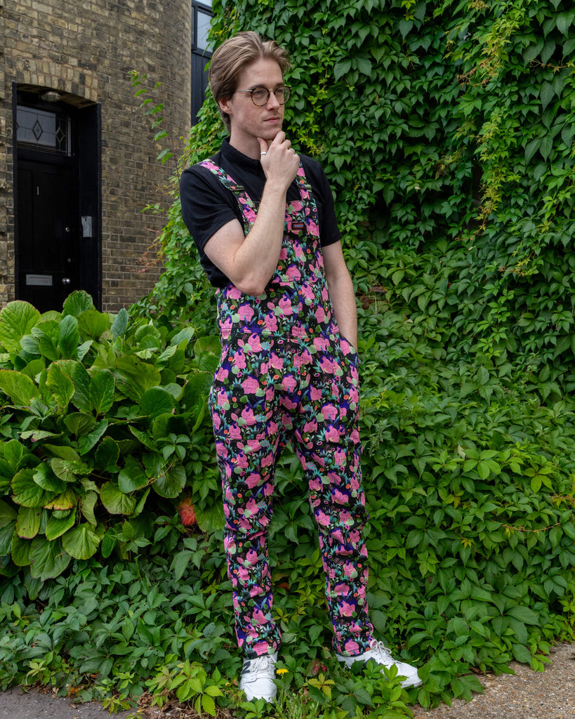 Model Jack Roper wears vibrant Run & Fly stretch twill dungarees featuring a fun 'Bodies' print by marie boiseau. The colourful pattern showcases pink bodies with green florals and fun details, paired with a black polo shirt. Set against lush greenery, this ethical slow fashion piece celebrates unique, sustainable style. Perfect for lovers of playful, bold fashion. Run & Fly is a small UK-based brand known for ethical, eco-conscious clothing.