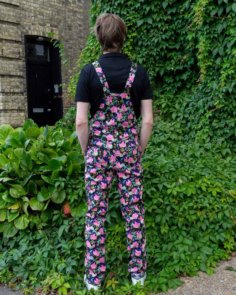 Person standing with their back to the camera, wearing Run & Fly dungarees with a colourful print featuring curvy, nude bodies and botanical patterns. The scene is set against a green ivy-covered wall, highlighting the slow fashion brand's ethical ethos. The black T-shirt and white trainers add contrast to the vibrant print, showcasing the playful design.