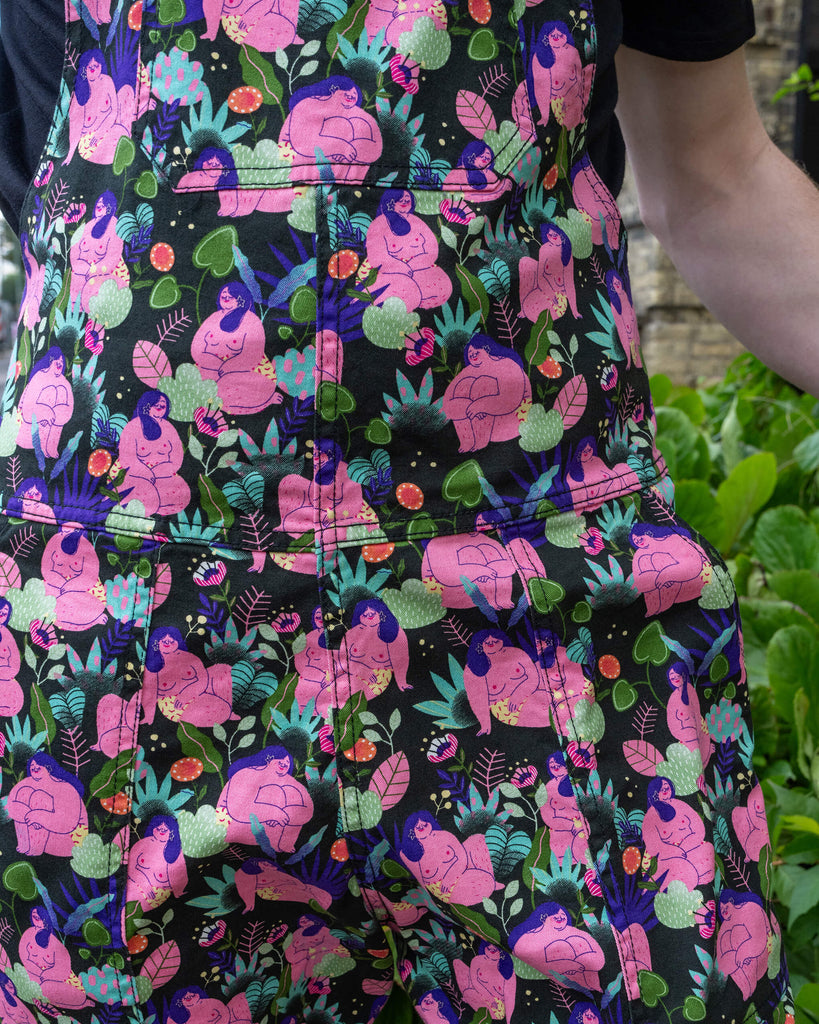 Detailed close-up of Run & Fly dungarees with a bold, artistic print of curvy nude bodies surrounded by plants and fungi. The vibrant pink, purple, and green hues contrast beautifully against the black fabric. These ethical dungarees are part of Run & Fly's slow fashion collection, offering unique and statement-making fashion.