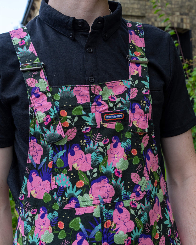 Close-up of a person wearing Run & Fly dungarees, featuring a bold print of curvy, nude figures with vibrant green plants and mushrooms. The pink, purple, and green colours stand out against a black background, while the person pairs it with a black collared shirt. This slow fashion brand offers ethical, fun, and statement clothing.