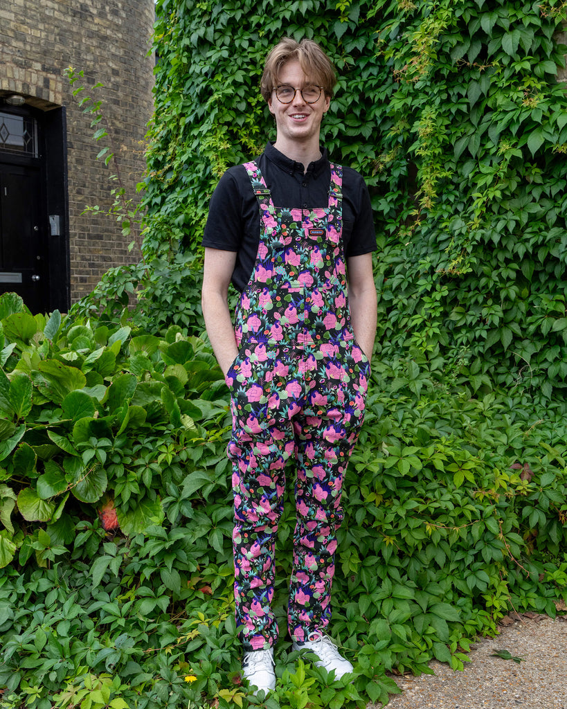 A smiling person in Run & Fly dungarees stands in front of a green ivy-covered wall. The dungarees feature an artistic print of nude, curvy bodies among plants, adding a playful and bold look. The white trainers and black shirt create a casual contrast to the colourful print, highlighting Run & Fly's ethical, slow fashion approach.