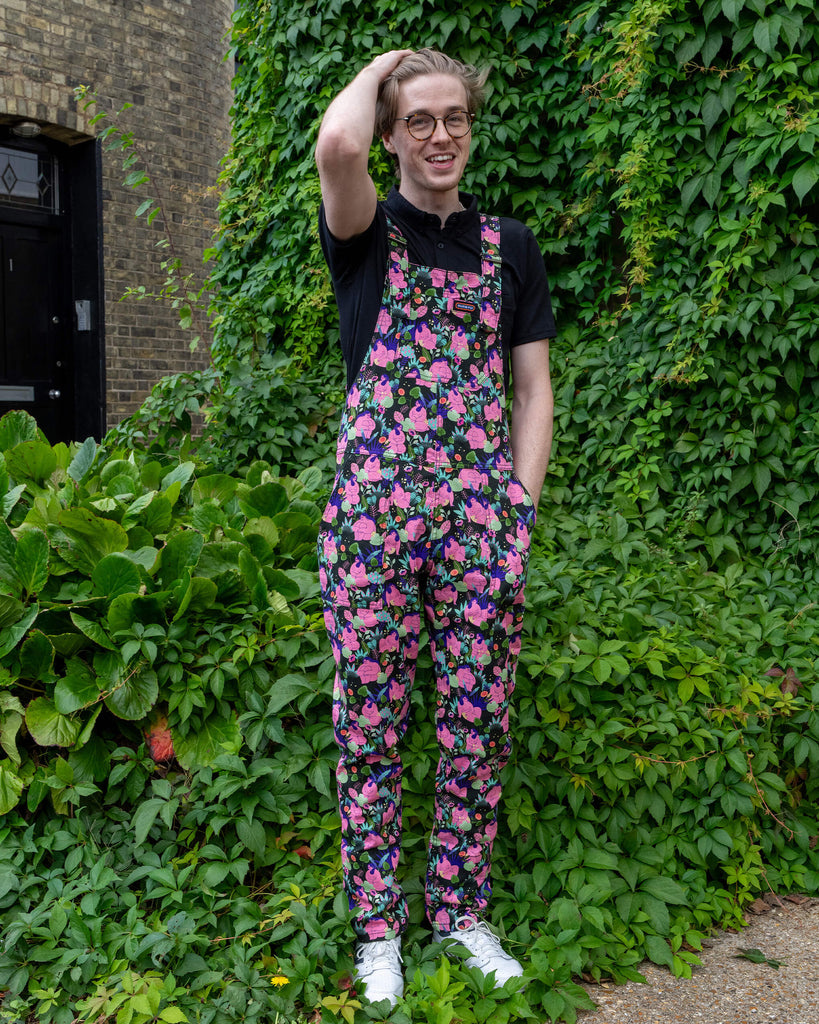 A smiling man with blonde hair, wearing glasses and a black polo shirt, poses outdoors in front of a lush green ivy wall. He is wearing vibrant, pink floral patterned dungarees by Run & Fly, a small ethical slow fashion brand. The bright pink flowers and green foliage on the black fabric stand out, paired with white trainers. The man stands with one hand on his head, looking relaxed and cheerful amidst the greenery.