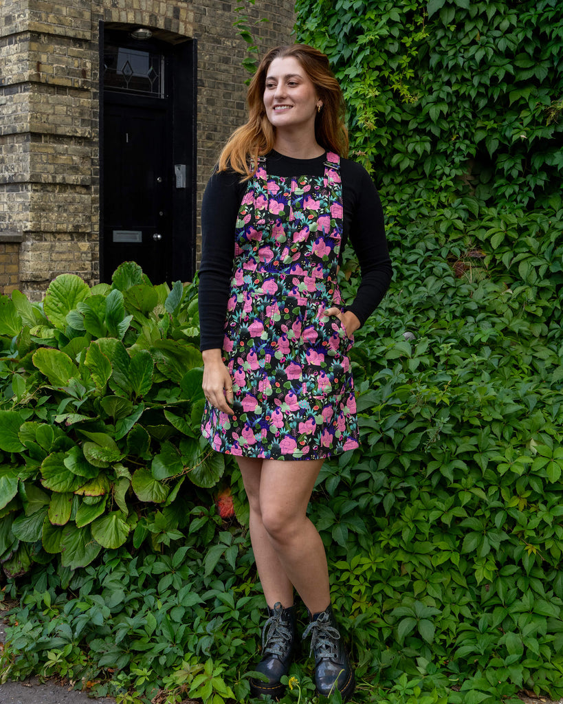 A woman models a vibrant floral pinafore dress by Run & Fly. She stands confidently in front of a leafy green background, wearing black boots and a black long-sleeve top. Run & Fly is a small ethical slow fashion brand.