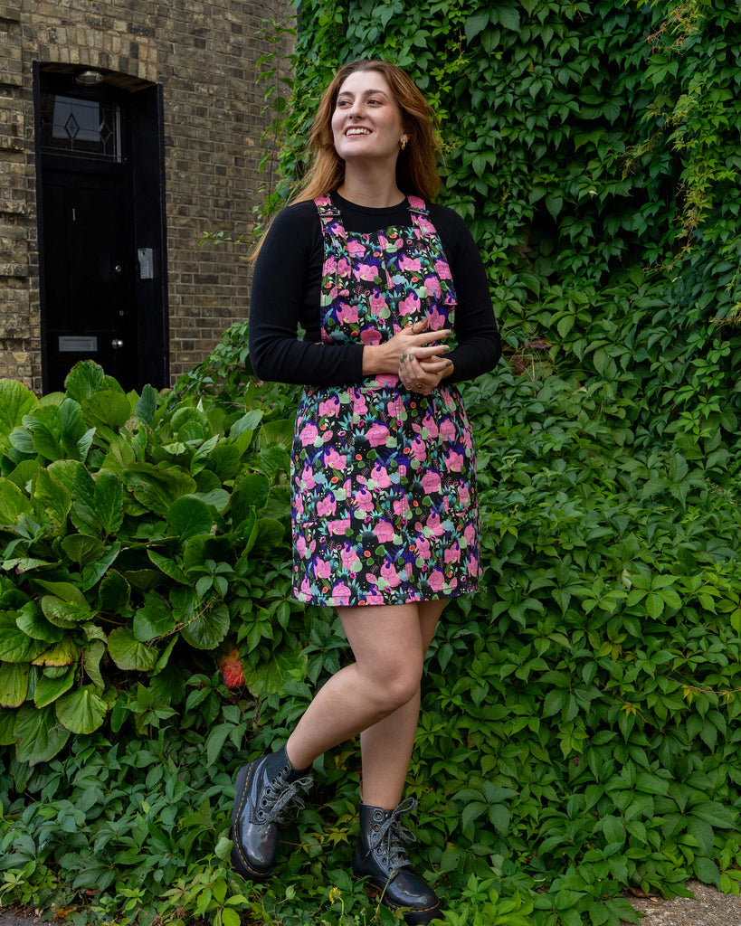 A woman models a floral pinafore dress by Run & Fly, standing confidently in front of lush greenery. She pairs the dress with black boots and a black long-sleeve top, smiling as she poses. Run & Fly is a small ethical slow fashion brand.