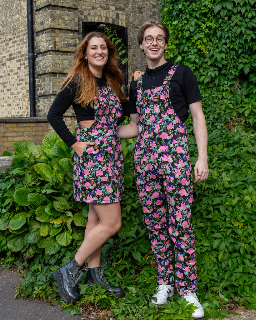 Two models wearing matching floral patterned outfits by Run & Fly, a small ethical slow fashion brand. The female model wears a pinafore dress paired with black boots, while the male model wears dungarees with white trainers. Both are standing outdoors in front of a lush green background and brick wall, smiling.