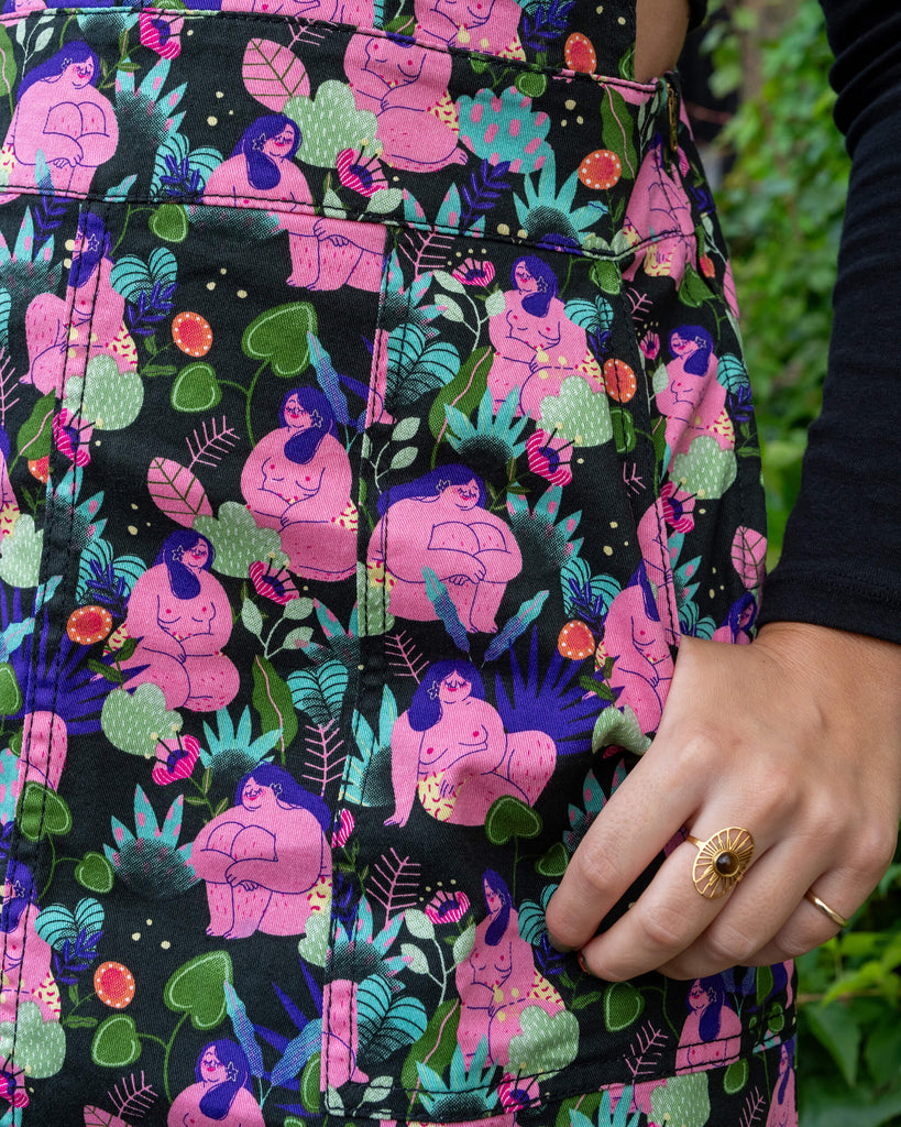 Close-up of the pocket detail on a floral pinafore dress by Run & Fly, showcasing a vibrant design of curvy, joyful figures among plants. The model's hand is tucked into the pocket, and she wears a gold ring. Run & Fly is a small ethical slow fashion company.