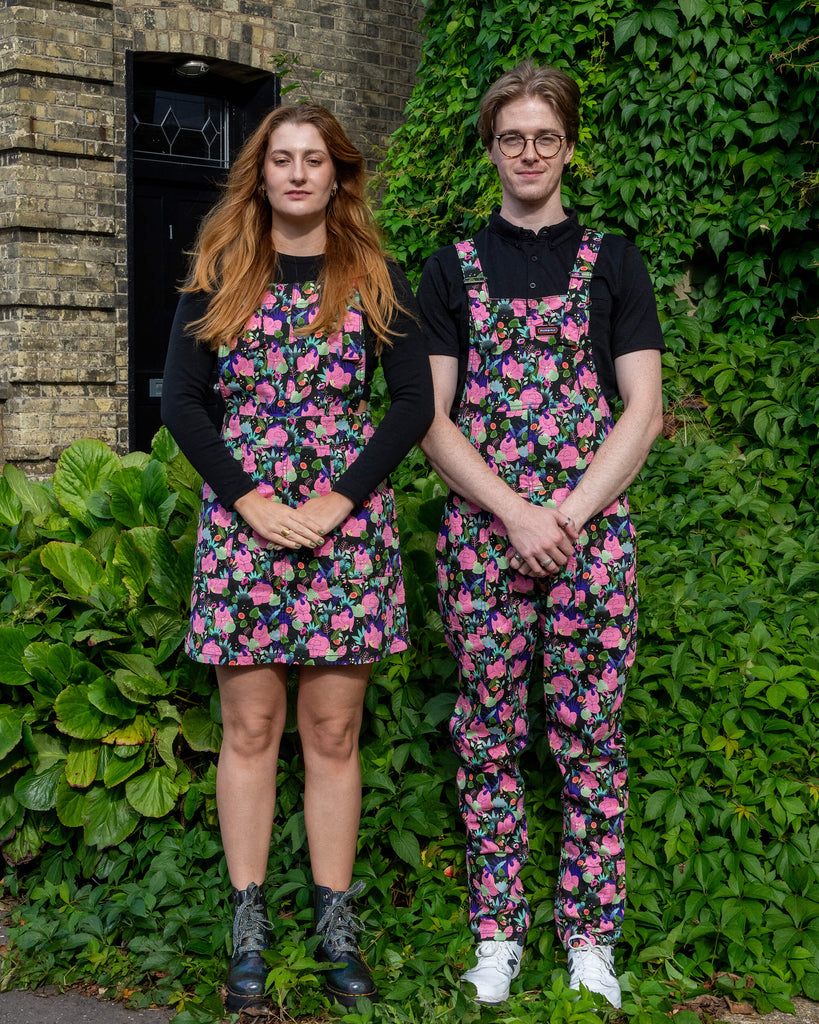 A woman and a man wearing matching floral outfits by Run & Fly, a small ethical slow fashion company. The woman wears a pinafore dress, and the man wears dungarees. They both stand with hands clasped in front, posing in front of a green background and a brick wall.