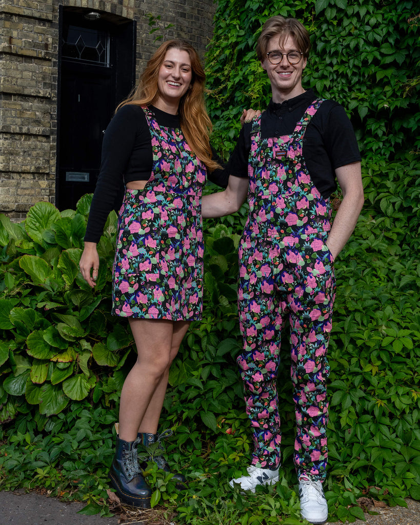 Two models in matching Run & Fly outfits, a small ethical slow fashion brand. The woman wears a floral pinafore dress, while the man wears dungarees with the same floral print. Both are standing in front of greenery, the woman in black boots, and the man in white trainers, smiling as they pose.