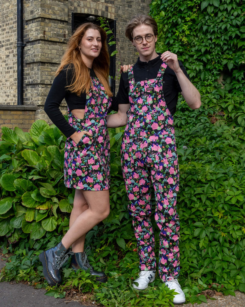 Two models wearing matching floral patterned outfits by Run & Fly, a small ethical slow fashion brand. The female model wears a pinafore dress paired with black boots, and the male model wears dungarees with white trainers. They are standing outdoors, posed against lush greenery and a brick wall, smiling.