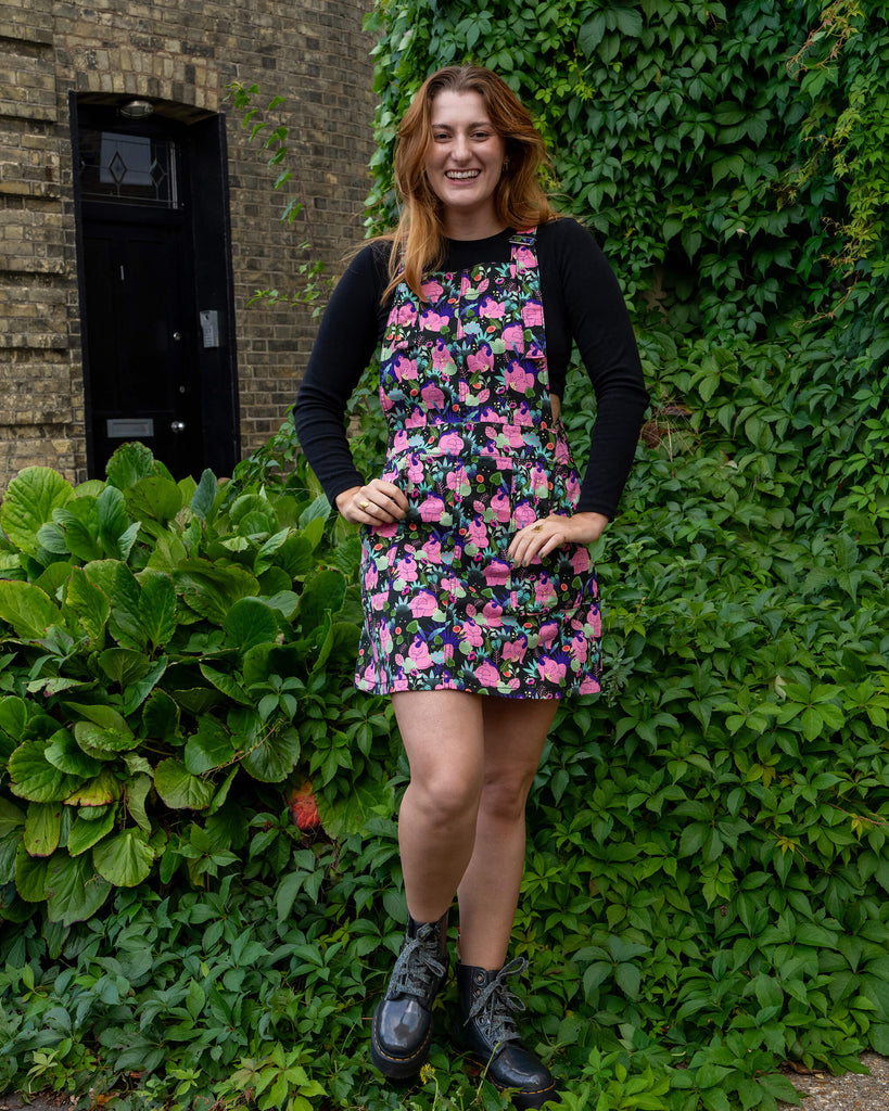 A woman smiles while modelling a floral pinafore dress by Run & Fly, standing in front of greenery and a brick building. She pairs the look with black boots and a black long-sleeve top. Run & Fly is a small ethical slow fashion company.
