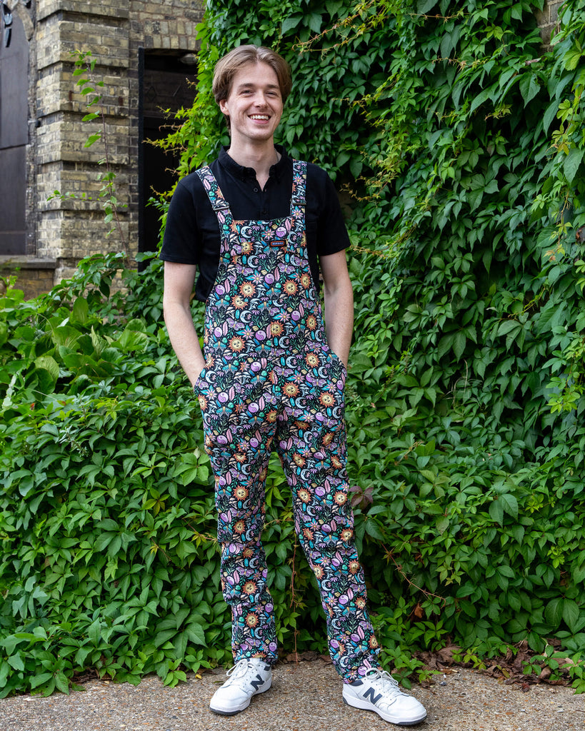 Jack Roper models colourful “Boho Summer” dungarees by Run & Fly, featuring a vibrant print with suns, moons, and flowers. Paired with a black shirt and white trainers, Jack smiles while posing in front of greenery, embodying the playful and ethical slow fashion ethos of the brand.