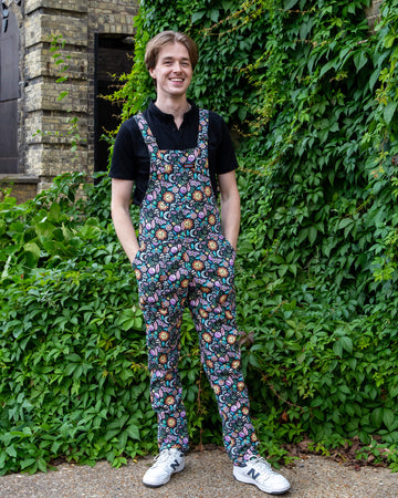 Jack Roper models colourful “Boho Summer” dungarees by Run & Fly, featuring a vibrant print with suns, moons, and flowers. Paired with a black shirt and white trainers, Jack smiles while posing in front of greenery, embodying the playful and ethical slow fashion ethos of the brand.