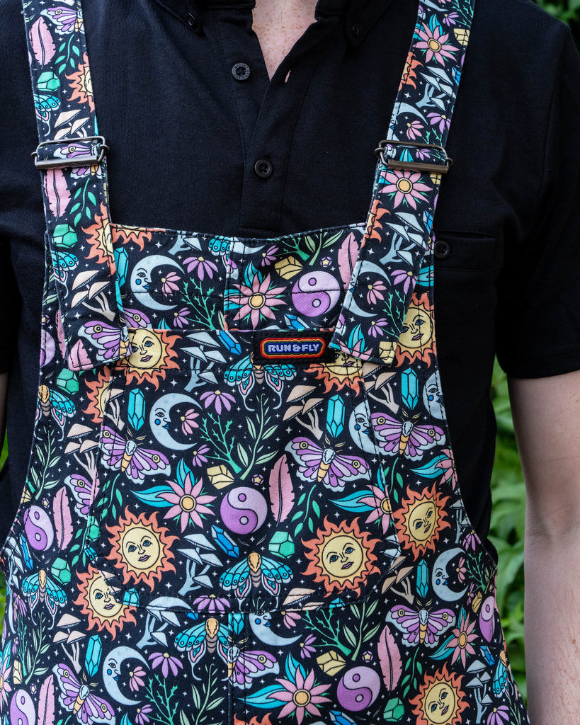 A close-up of Jack Roper wearing Run & Fly’s “Boho Summer” dungarees, focusing on the vibrant print featuring celestial suns, moons, and floral elements. The black shirt underneath complements the detailed, colourful design.