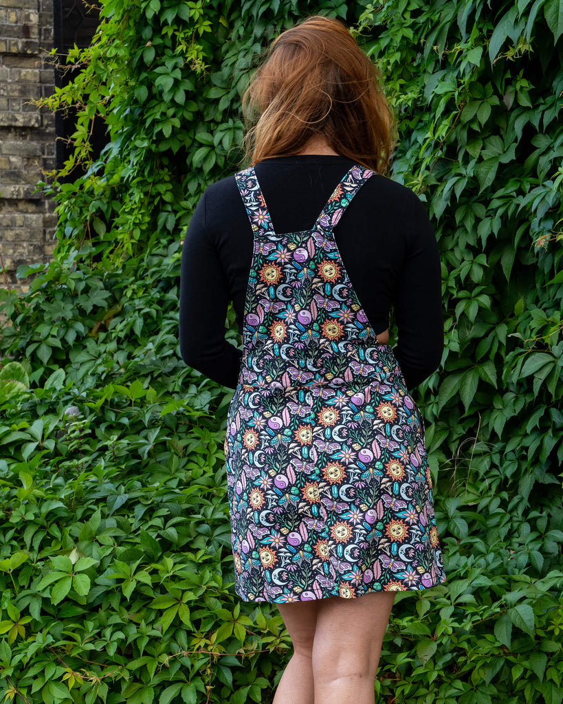 Katie is stood outside in Hove by greenery wearing Run & Fly Boho Summer Stretch Twill Pinafore Dress with a long sleeved black top underneath and white trainers. The pinafore has a dark base with an all over print of flowers, moths, flowers, suns and more. She is stood with her back to the camera with one leg slightly bent.