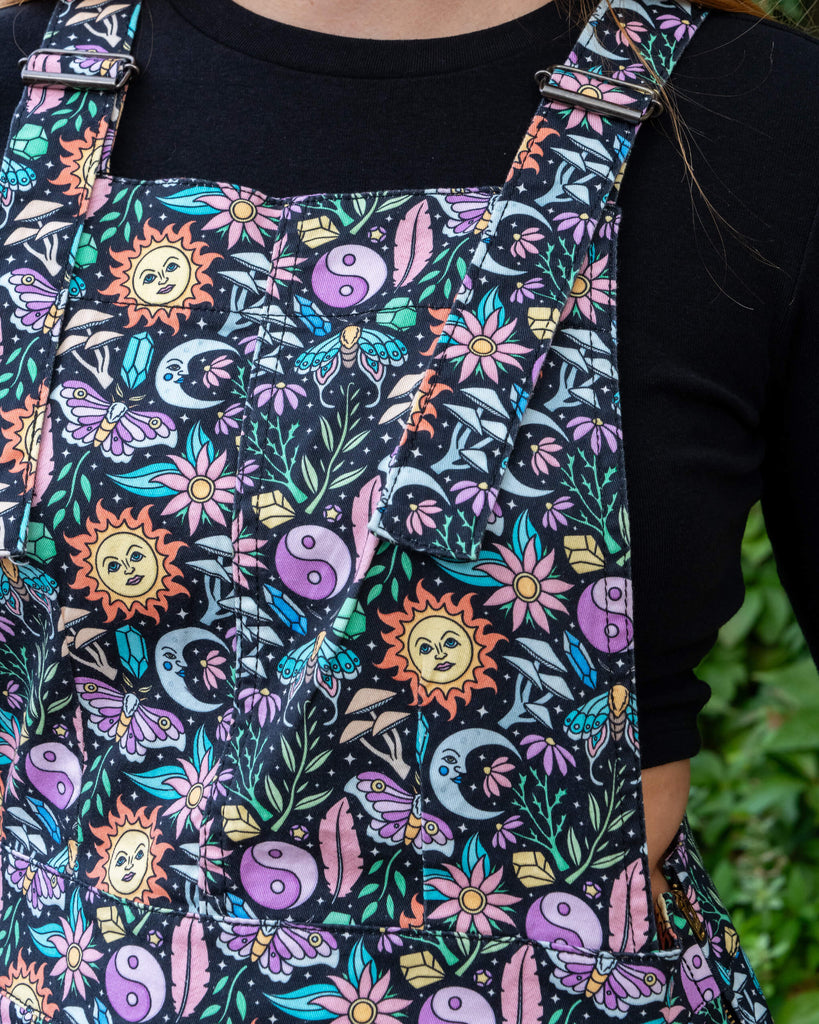 Close up of model wearing Run & Fly Boho Summer Stretch Twill Pinafore Dress. The pinafore has a dark base with an all over print of flowers, moths, flowers, suns and more.