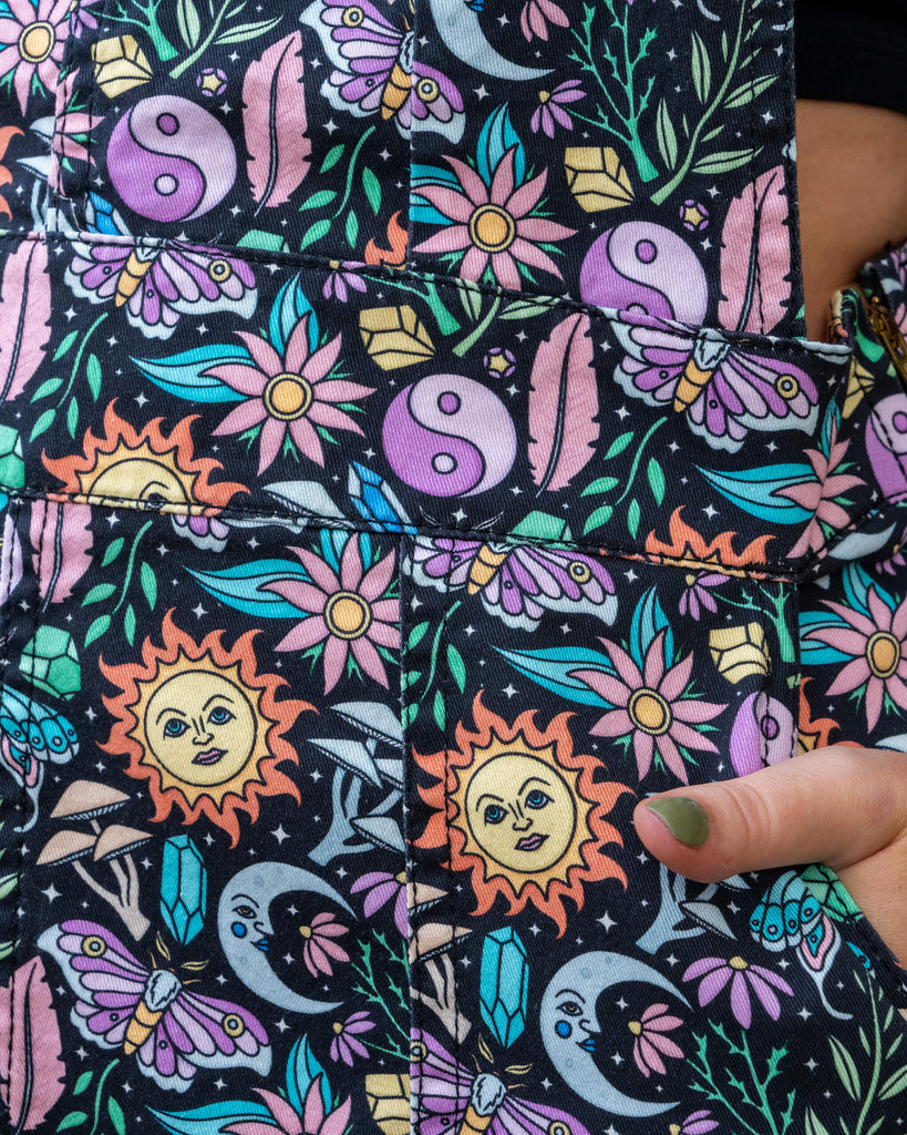 Close up of model wearing Run & Fly Boho Summer Stretch Twill Pinafore Dress. The pinafore has a dark base with an all over print of flowers, moths, flowers, suns and more. Close up shows model's hand in the pocket of the pinafore. 