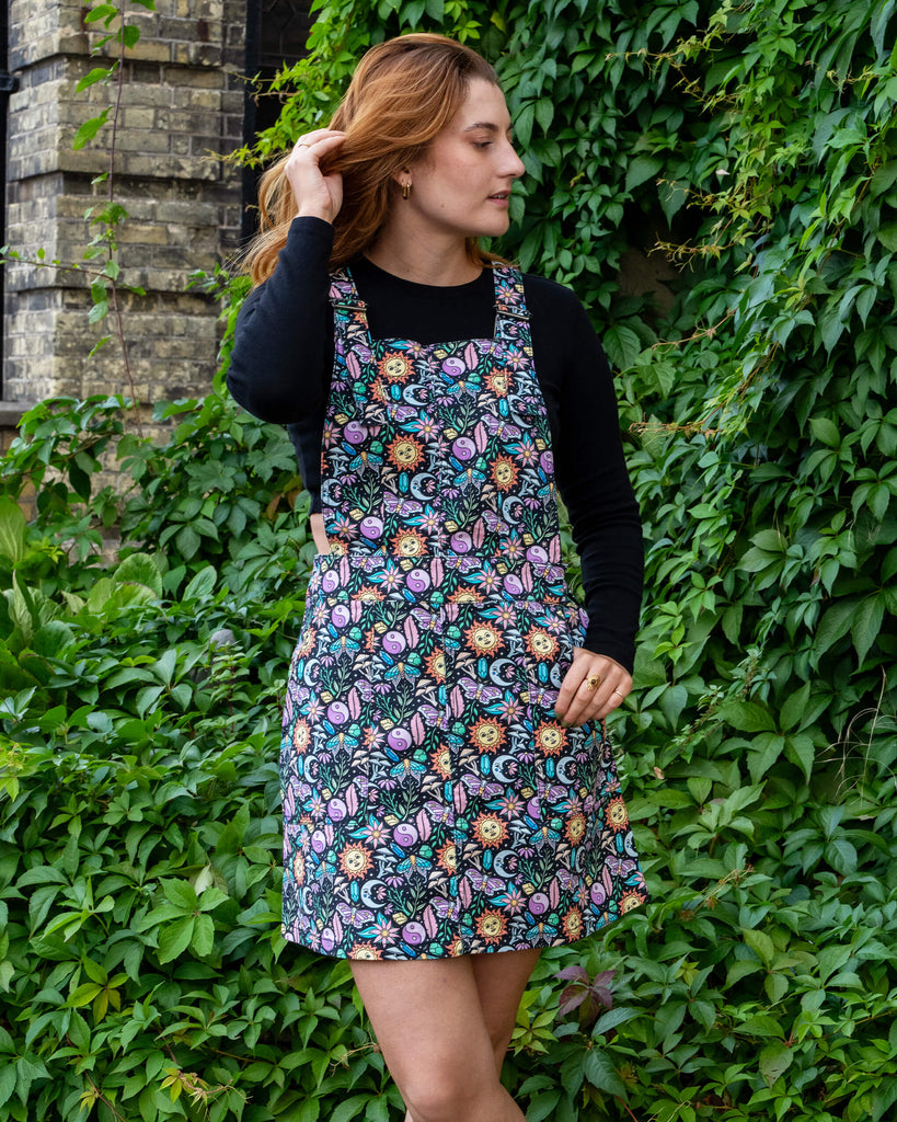 Katie is stood outside in Hove by greenery wearing Run & Fly Boho Summer Stretch Twill Pinafore Dress with a long sleeved black top underneath. The pinafore has a dark base with an all over print of flowers, moths, flowers, suns and more. She is posing with one hand resting in the pinafore pocket and is looking off to one side with one leg in front of the other.