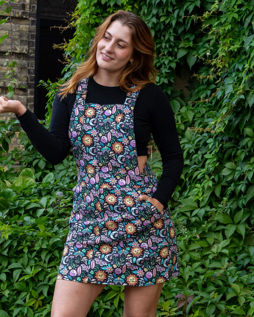 Katie is stood outside in Hove by greenery wearing Run & Fly Boho Summer Stretch Twill Pinafore Dress with a long sleeved black top underneath. The pinafore has a dark base with an all over print of flowers, moths, flowers, suns and more. She is posing with one hand resting in the pinafore pocket and is looking off to one side with one leg in front of the other.