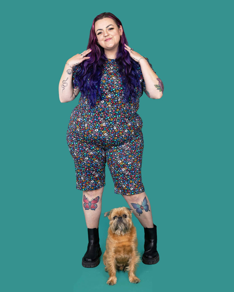 The Boogie Bones Short Sleeve T Shirt worn by a femme alternative tattooed model with long pink and purple hair and the matching shorts on a teal studio background. She is facing forward smiling with both arms resting by her sides. The black base tshirt features dancing smiling skeletons with multicoloured flowers in various sizes and types all over.