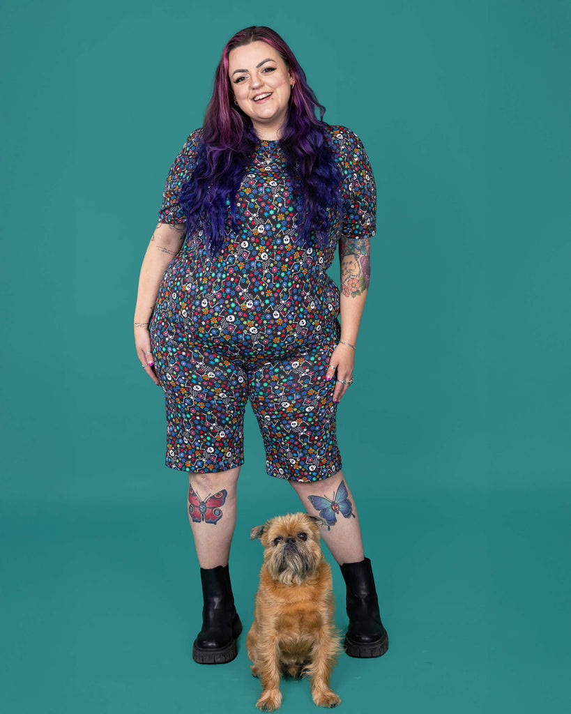 The Boogie Bones Stretch Cycle Shorts worn by a femme alternative tattooed model with long pink and purple hair, the matching tshirt and black boots on a teal studio background with a small brown dog. She is facing forward laughing with both hands resting by her sides. The black base shorts features dancing smiling skeletons with multicoloured flowers in various sizes and types all over.