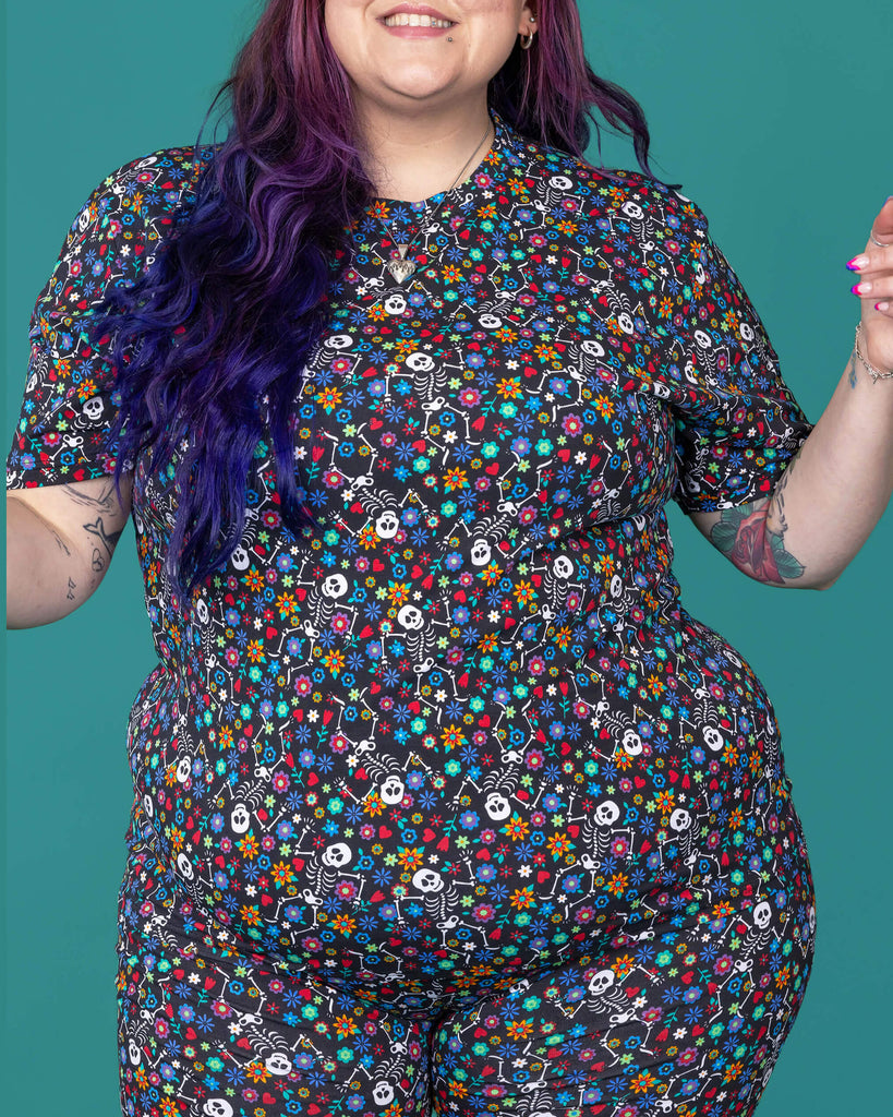 The Boogie Bones Short Sleeve T Shirt worn by a femme alternative tattooed model with long pink and purple hair and the matching shorts on a teal studio background. She is facing forward smiling with both arms resting by her sides. The black base tshirt features dancing smiling skeletons with multicoloured flowers in various sizes and types all over.