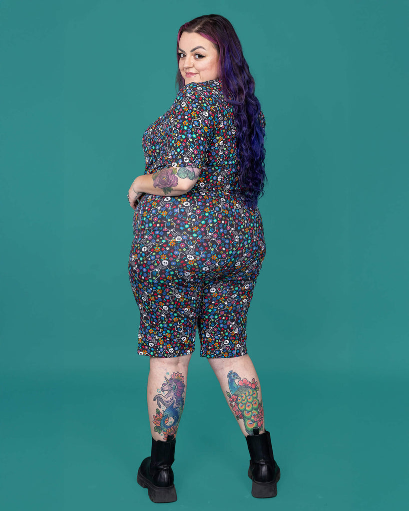 The Boogie Bones Stretch Cycle Shorts worn by a femme alternative tattooed model with long pink and purple hair, the matching tshirt and black boots on a teal studio background. She is facing away smiling looking over her left shoulder with her arms resting on her hips. The black base shorts features dancing smiling skeletons with multicoloured flowers in various sizes and types all over.
