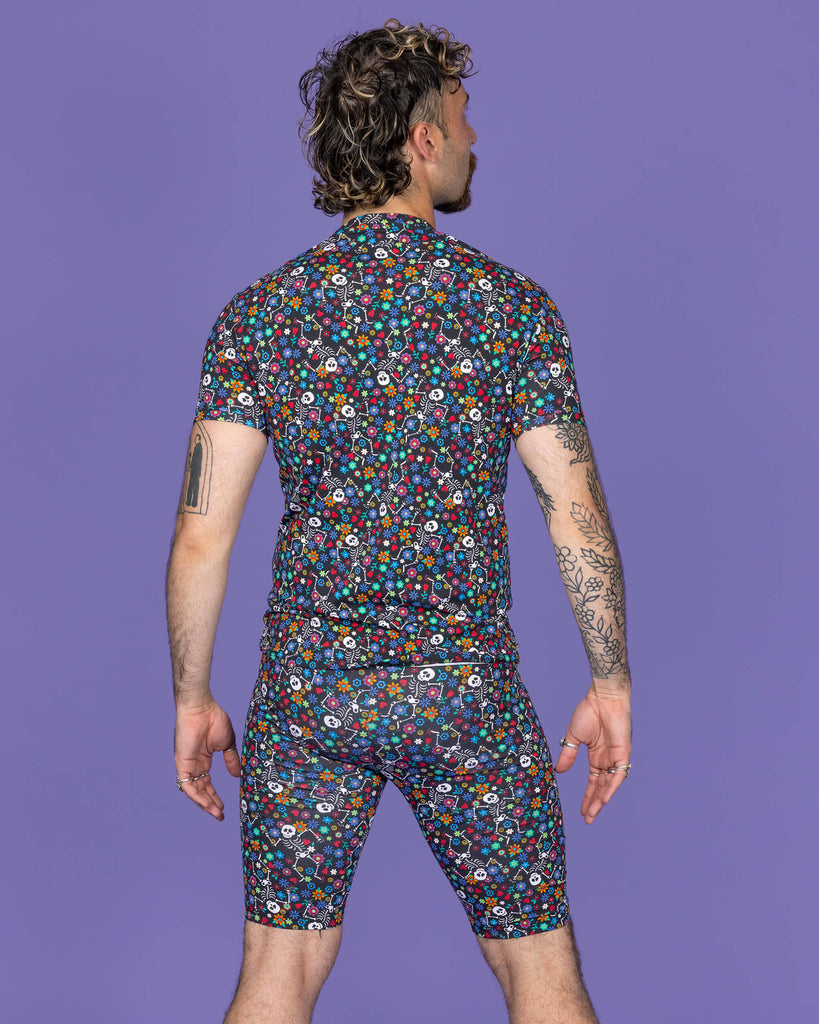The Boogie Bones Stretch Cycle Shorts worn by a masc non binary model with a brown curly mullet, the matching tshirt and white trainers on a purple studio background. They are facing away from camera looking off to the right with both arms resting by their sides. The black base shorts features dancing smiling skeletons with multicoloured flowers in various sizes and types all over.