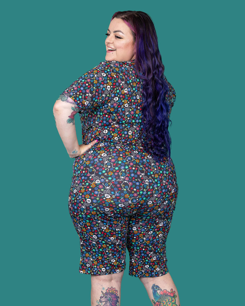 The Boogie Bones Short Sleeve T Shirt worn by a femme alternative tattooed model with long pink and purple hair and the matching shorts on a teal studio background. She is facing away laughing looking over her left shoulder with her hands on her hips. The black base tshirt features dancing smiling skeletons with multicoloured flowers in various sizes and types all over.