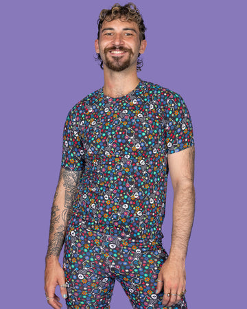 The Boogie Bones Short Sleeve T Shirt worn by a masc non binary model with a brown curly mullet, the matching cycle shorts on a purple studio background. They are facing forward smiling leaning more on their left hip with both hands resting by their sides. The black base tshirt features dancing smiling skeletons with multicoloured flowers in various sizes and types all over.