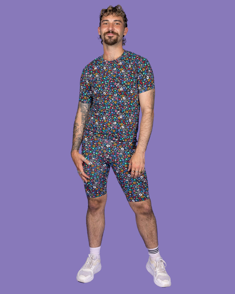 The Boogie Bones Short Sleeve T Shirt worn by a masc non binary model with a brown curly mullet, the matching cycle shorts and white trainers on a purple studio background. They are facing forward smiling leaning more on their left hip with both hands resting by their sides. The black base tshirt features dancing smiling skeletons with multicoloured flowers in various sizes and types all over.