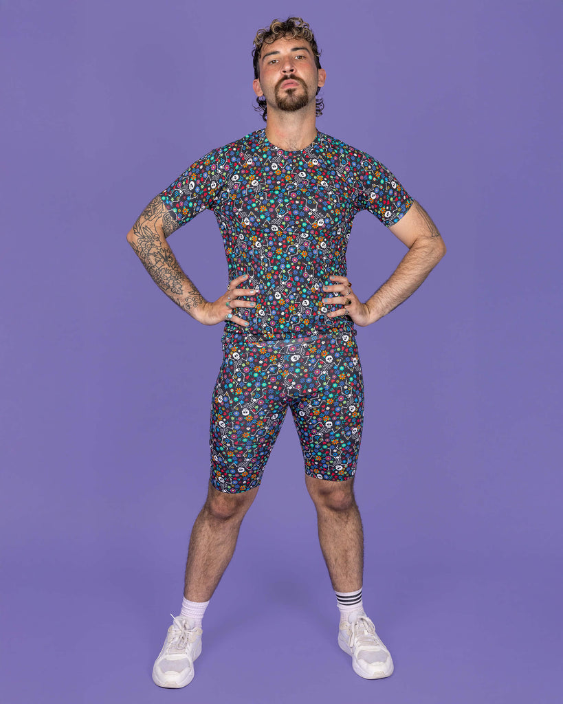 The Boogie Bones Stretch Cycle Shorts worn by a masc non binary model with a brown curly mullet, the matching tshirt and white trainers on a purple studio background. They are facing forward with their head tilted slightly back looking to camera with both hands on their hips. The black base shorts features dancing smiling skeletons with multicoloured flowers in various sizes and types all over.