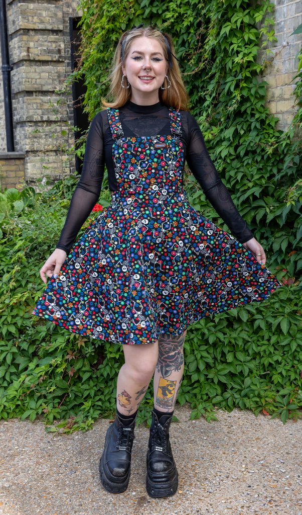 Florence, a white female model with blonde hair and tattoos is stood outside in Hove wearing Halloween: Boogie Bones Flared Pinafore Dress with a long sleeved mesh black top underneath and chunky black boots. Boogie bones print is a black base with multicoloured flowers and dancing smiling cartoon skeletons all over. Florence is smiling toward the camera holding the flared skirt of the dress.