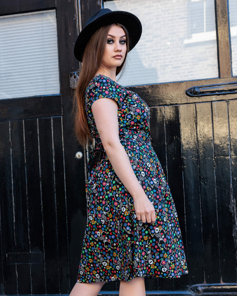 Charlotte wearing the Halloween: Boogie Bones Stretch Belted Tea Dress with Pockets a witchy styled wide brim black hat. She is facing away and looking over her right shoulder to camera with one hand on her hip and the other resting by her side. Charlotte has long brown hair and dark smokey makeup. Boogie bones print is a black base with multicoloured flowers and dancing smiling cartoon skeletons all over, the dress is short sleeved, round neck and has a belt tie.
