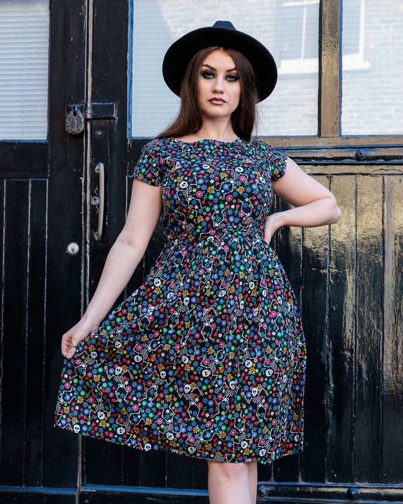 Charlotte wearing the Halloween: Boogie Bones Stretch Belted Tea Dress with Pockets with a witchy styled wide brim black hat. She is facing forward with one hand on her hip and the other holding out the bottom of the dress. Charlotte has long brown hair and dark smokey makeup. Boogie bones print is a black base with multicoloured flowers and dancing smiling cartoon skeletons all over, the dress is short sleeved, round neck and has a belt tie.