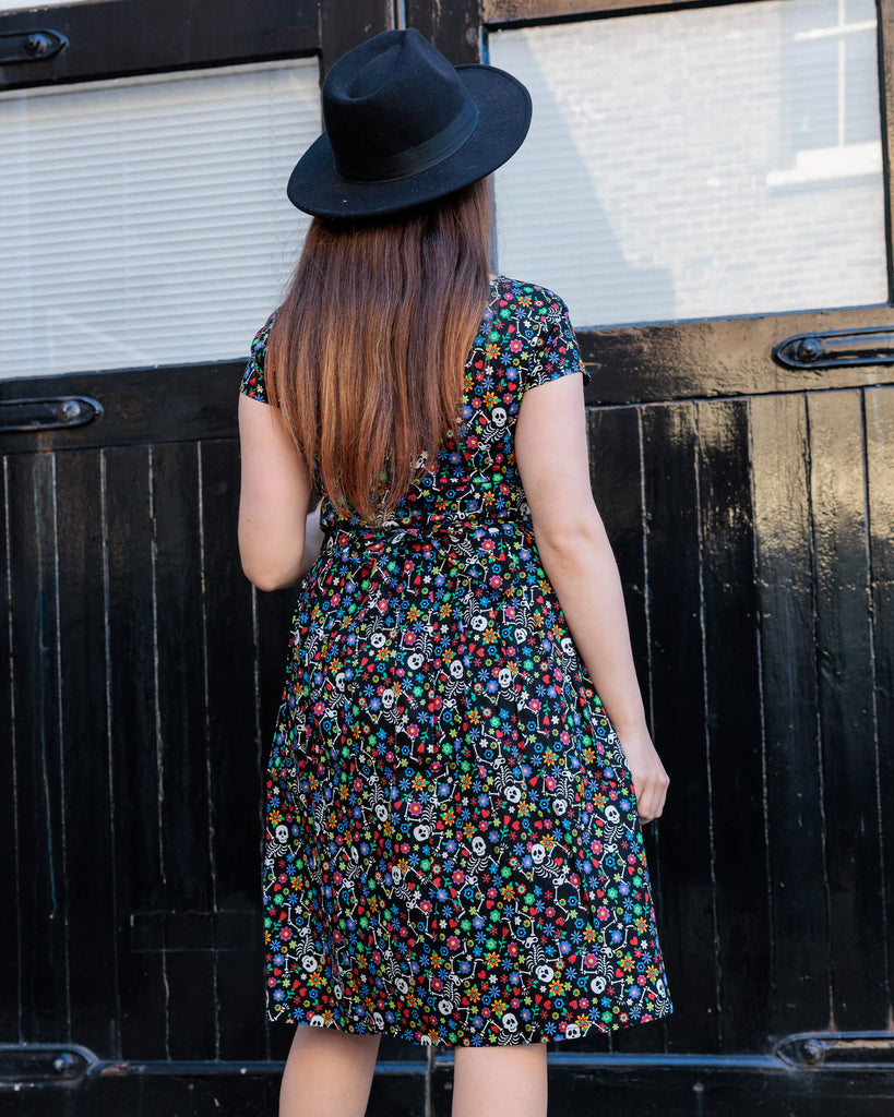 Charlotte wearing the Halloween: Boogie Bones Stretch Belted Tea Dress with Pockets with a witchy styled wide brim black hat. She is facing away from the camera with one hand on her hip and the other resting by her side. Charlotte has long brown hair and dark smokey makeup. Boogie bones print is a black base with multicoloured flowers and dancing smiling cartoon skeletons all over, the dress is short sleeved, round neck and has a belt tie.