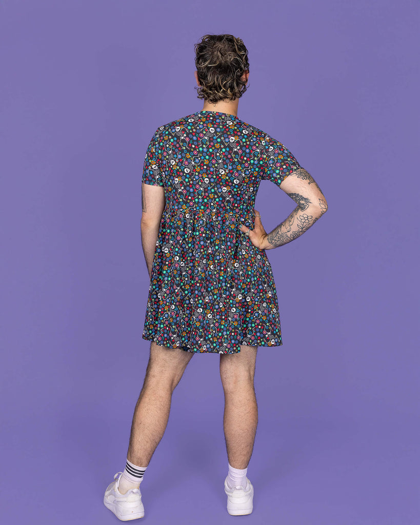 The Boogie Bones Oversized Smock Dress on a masc non binary model with a short curly brown mullet and white trainers on a purple studio background. They are facing away leaning more on their right hip with their right hand on their hip. The black base dress features dancing smiling skeletons with multicoloured flowers in various sizes and types all over.