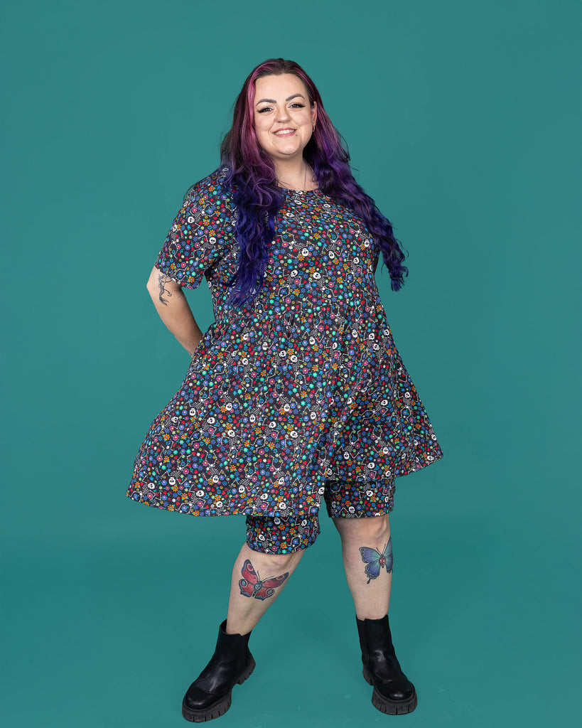 The Boogie Bones Oversized Smock Dress worn by a femme alternative tattooed model with long pink and purple hair, the matching shorts and black boots on a teal studio background. She is facing forward smiling with her hands behind her back twirling the dress out. The black base dress features dancing smiling skeletons with multicoloured flowers in various sizes and types all over.