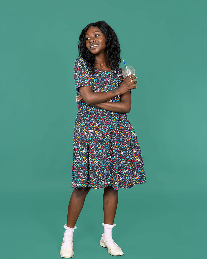 The Boogie Bones Oversized Smock Dress worn by a femme alternative model with mid length black hair with white frilly socks, white dolly style heeled shoes and holding an iced coffee on a teal studio background. She is facing forward with her arms folded across her front holding an iced coffee and smiling looking left. The black base dress features dancing smiling skeletons with multicoloured flowers in various sizes and types all over.
