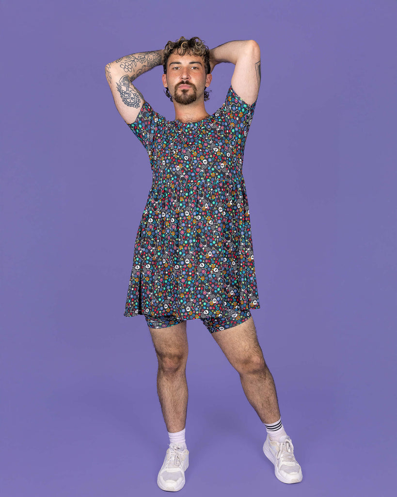 The Boogie Bones Oversized Smock Dress on a masc non binary model with a short curly brown mullet and white trainers on a purple studio background. They are facing forward with their arms resting behind their head whilst pouting to camera. The black base dress features dancing smiling skeletons with multicoloured flowers in various sizes and types all over.