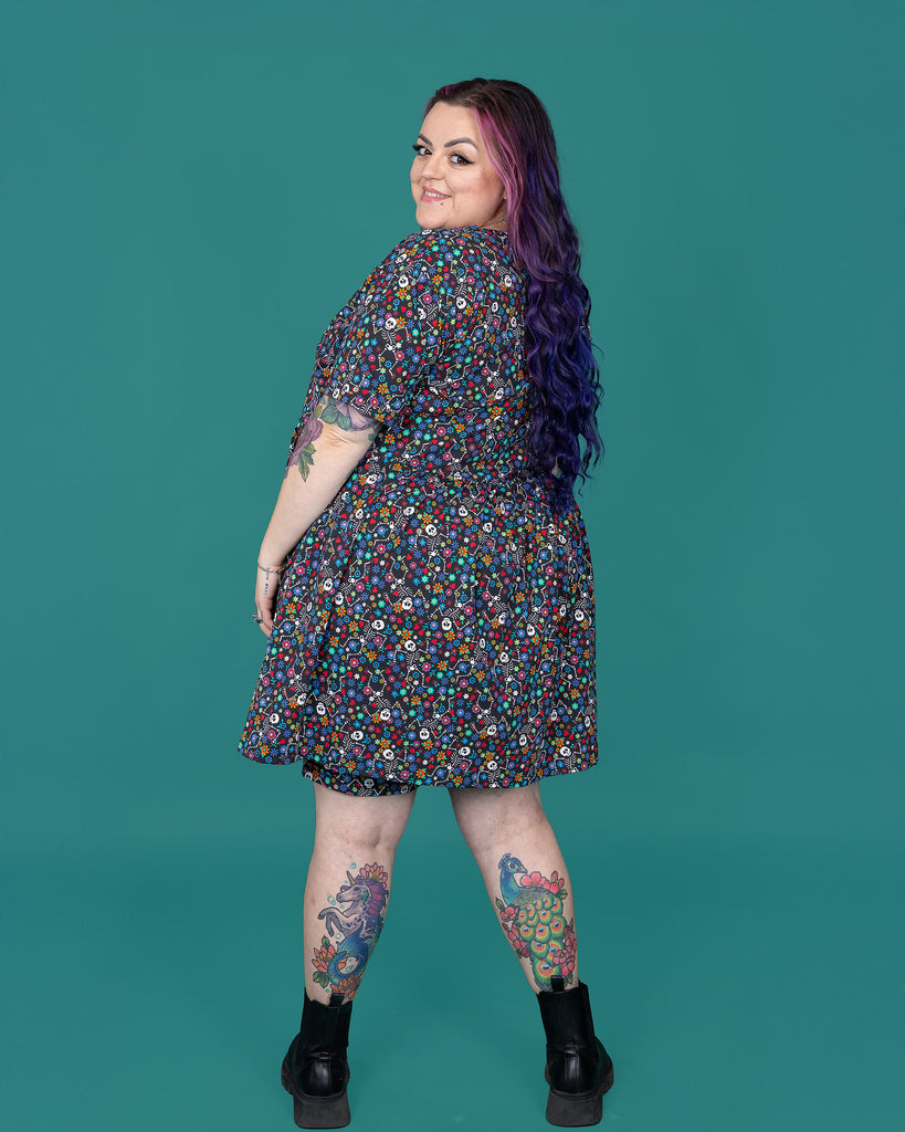 The Boogie Bones Oversized Smock Dress worn by a femme alternative tattooed model with long pink and purple hair, the matching shorts and black boots on a teal studio background. She is facing away smiling looking over her left shoulder with her arms resting by her sides. The black base dress features dancing smiling skeletons with multicoloured flowers in various sizes and types all over.