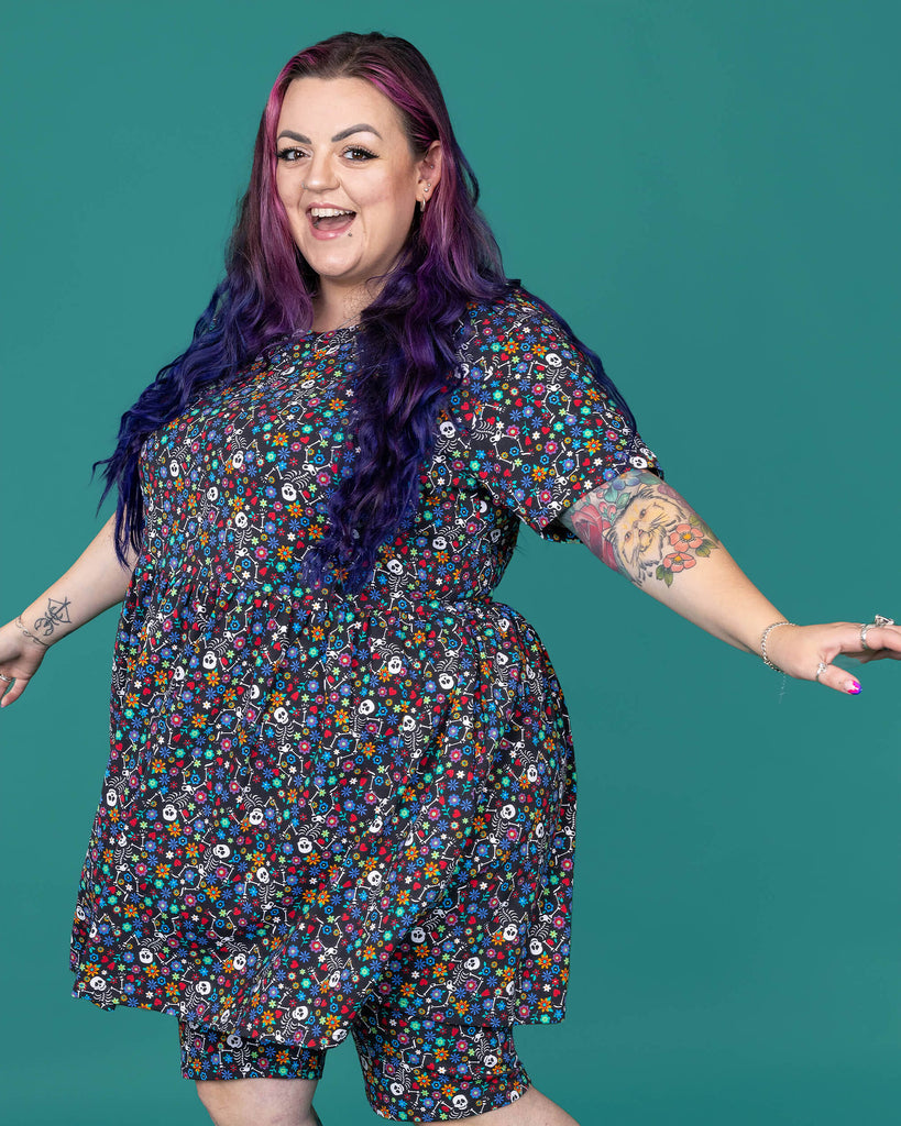 The Boogie Bones Oversized Smock Dress worn by a femme alternative tattooed model with long pink and purple hair, the matching shorts and black boots on a teal studio background. She is taking a step to the left laughing with her hands out to the side. The black base smock dress features dancing smiling skeletons with multicoloured flowers in various sizes and types all over.