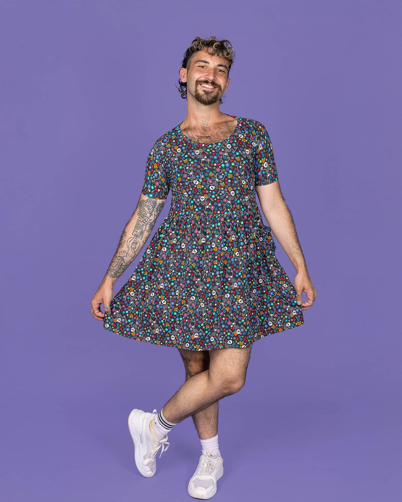 The Boogie Bones Oversized Smock Dress on a masc non binary model with a short curly brown mullet and white trainers on a purple studio background. They are facing forward smiling holding out the bottom of the skirt with one leg crossed over the other. The black base smock dress features dancing smiling skeletons with multicoloured flowers in various sizes and types all over.