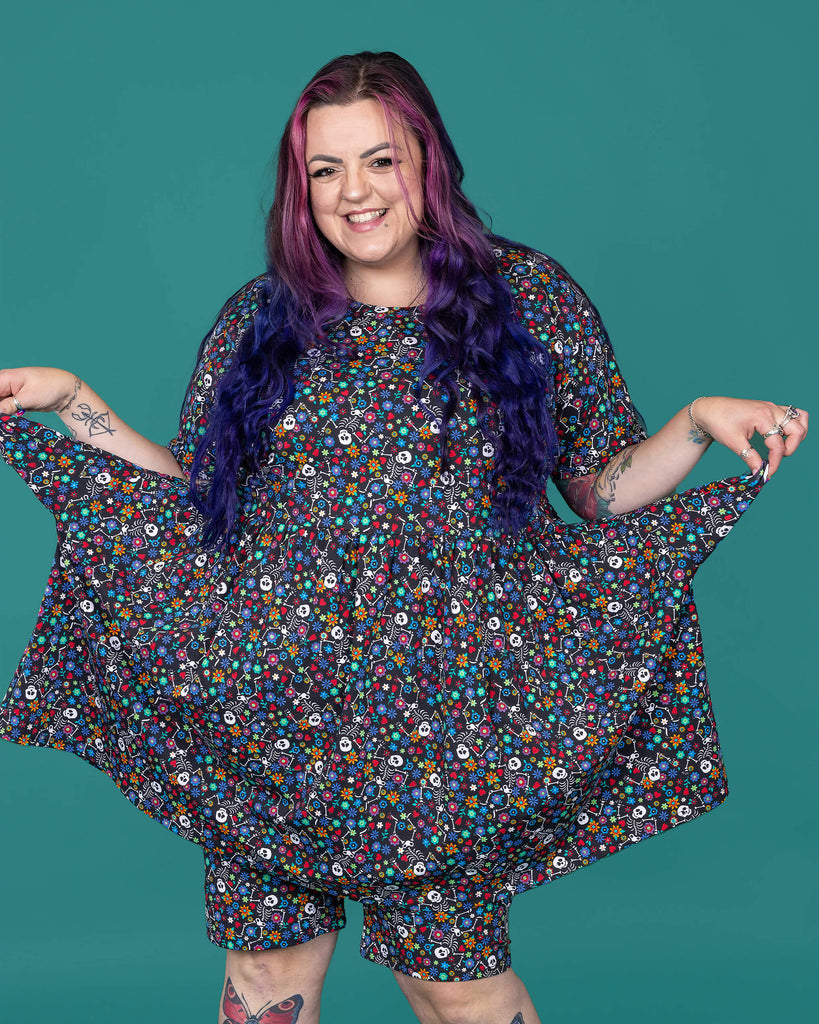 The Boogie Bones Oversized Smock Dress worn by a femme alternative tattooed model with long pink and purple hair, the matching shorts and black boots on a teal studio background. She is facing forward laughing holding out the dress to the sides by the pockets. The black base smock dress features dancing smiling skeletons with multicoloured flowers in various sizes and types all over.
